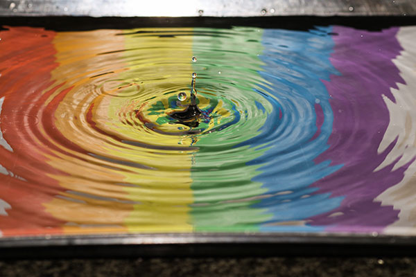 A water droplet hitting a pool of water, which is coloured in stripes, one for each colour of the rainbow